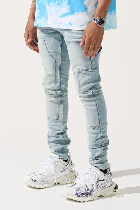 A person standing wearing light blue denim jeans with carpenter panels and a carpenter loop on the left leg, paired with a tie-dye shirt and white sneakers.